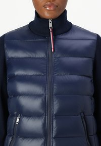 Gilet di piumino blu navy con alto collo, caratterizzato da sezioni trapuntate, due tasche laterali con zip e una chiusura lampo argentata con accento rosso.