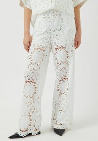 Pantaloni bianchi in pizzo con motivi floreali, vestibilità ampia e design a gamba larga. Abbinati a scarpe nere appuntite, che mostrano dettagli e texture raffinati.