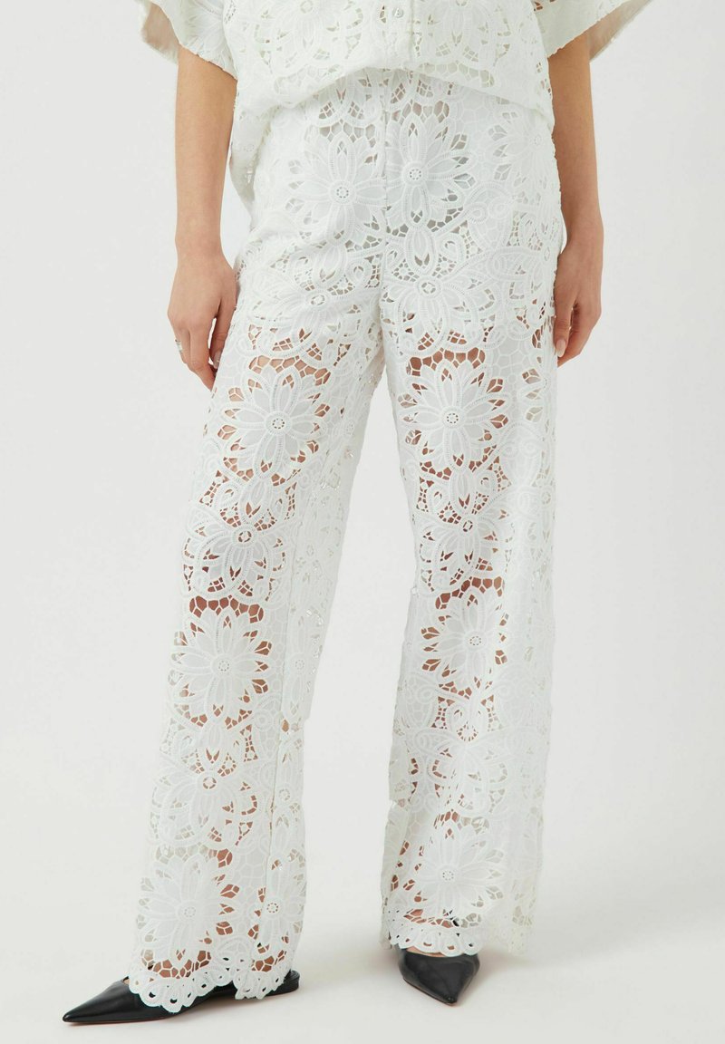 Pantaloni bianchi in pizzo con motivi floreali, vestibilità ampia e design a gamba larga. Abbinati a scarpe nere appuntite, che mostrano dettagli e texture raffinati.
