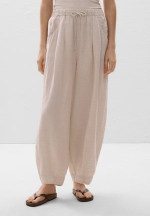Pantaloni in lino color beige chiaro con vita regolabile, pieghe e tasche laterali, abbinati a sandali infradito marrone scuro.