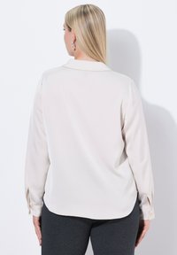 Blusa beige chiaro con colletto classico, maniche lunghe e polsini con bottoni. Texture liscia e lucida con un orlo leggermente curvo.