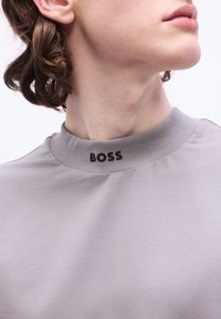 Άτομο με σγουρά μαλλιά που φοράει ανοιχτό γκρι πουλόβερ με ψηλό γιακά, όπου είναι τυπωμένο το "BOSS" με μαύρα γράμματα στο γιακά, το κεφάλι γυρισμένο πλάγια.