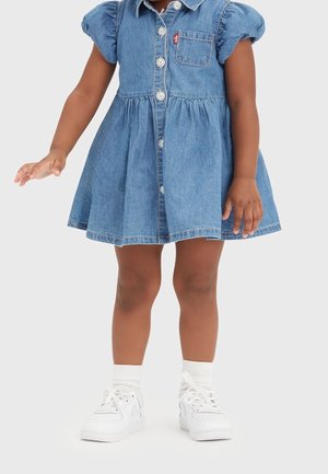 Dziecko w niebieskiej, krótko rękawowej sukience z dżinsu z guzikami, białych skarpetkach i białych tenisówkach, stojące na jednolitym tle.