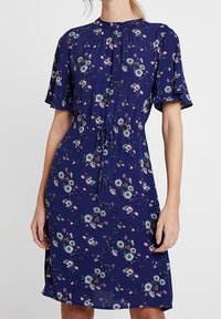 Navyblå blommig klänning med korta vågiga ärmar, dragsko i midjan och knä-lång nederdel. Har ett subtilt mönster av blommor och gröna växter.