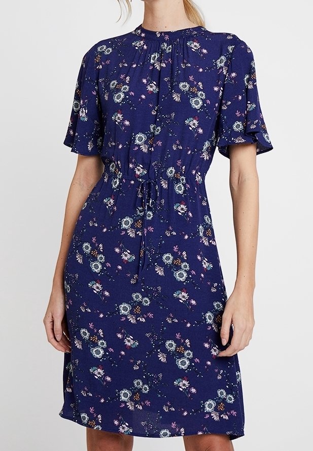 Navyblå blommig klänning med korta vågiga ärmar, dragsko i midjan och knä-lång nederdel. Har ett subtilt mönster av blommor och gröna växter.