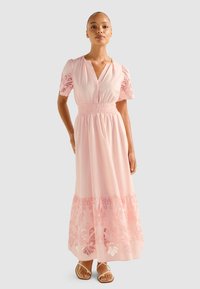 Rochie maxi din material roz deschis, cu mâneci scurte tip puff, decolteu în V, talie elastică și detaliu cu broderie florală pe tiv. Sandale cu cureluse subțiri.