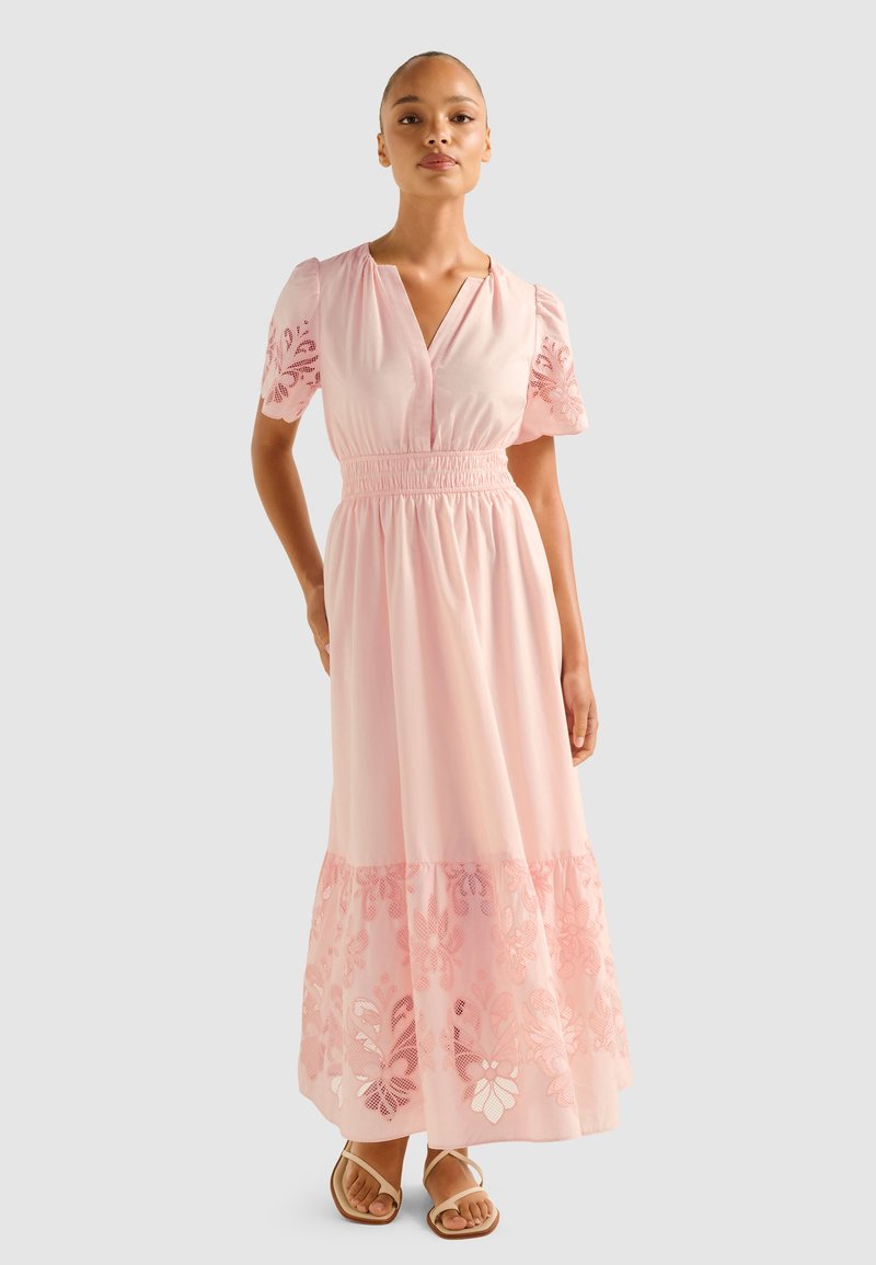 Rochie maxi din material roz deschis, cu mâneci scurte tip puff, decolteu în V, talie elastică și detaliu cu broderie florală pe tiv. Sandale cu cureluse subțiri.