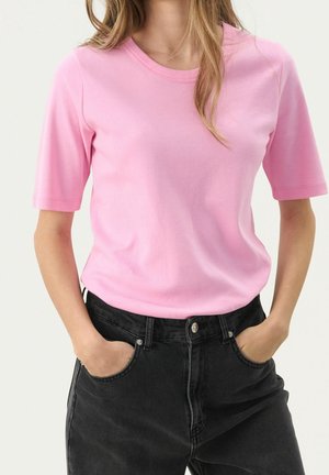 Vrouw draagt een effen roze shirt met korte mouwen, ingestopt in hooggesloten zwarte spijkerbroek, met handen in de zakken.