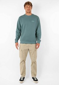 Un sweatshirt crewneck teal avec des poignets côtelés, associé à un pantalon cargo beige avec des poches latérales, et des baskets noires avec des semelles blanches.