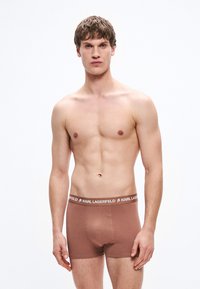 Καφέ βαμβακερά μποξεράκια με ελαστική μέση με λογότυπο, που προσφέρουν εφαρμογή που αγκαλιάζει το σώμα και απαλή υφή. Το μοντέλο στέκεται μπροστά σε λευκό φόντο.