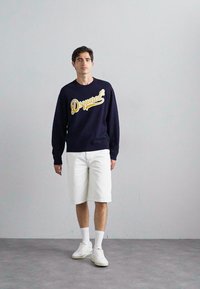 Tengerészsapkás pulóver sárga "Dsquared2" logóval, fehér nadrággal, fehér sportcipőben és zokniban áll egy világosszürke háttér előtt.