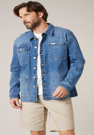 Man die een lichtblauwe denimjas met knopen en meerdere zakken draagt, samen met beige korte broek, kijkt opzij tegen een effen achtergrond.