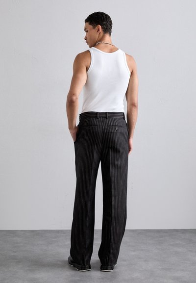 Black pinstriped trousers with a straight cut, made of a textured fabric. Worn with a white sleeveless shirt, featuring back pockets and a belt.