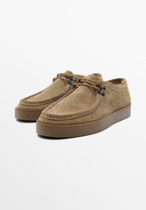 Braune Wildleder-Slipper mit abgerundeter Spitze, kontrastierenden Nähten und geflochtenen Schnürsenkeln. Weiches Innenfutter und flache Gummisohle.