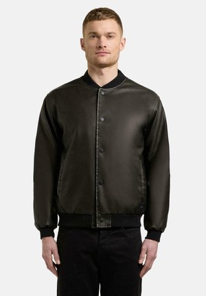 Mand iført en knappet sort læder bomberjakke med ribbet krave, manchetter og kant, stående mod en ensfarvet lys baggrund.