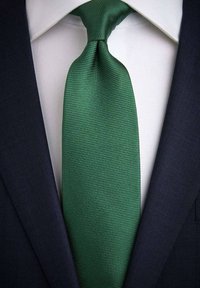 Grön texturerad slips med smal väv, bärs över en vit skjorta och mörk marinblå kostymjacka. Har en slät yta och klassisk knut.