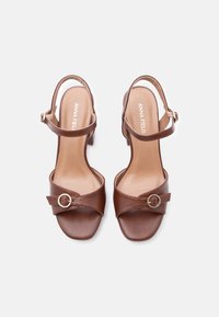 Καφέ δερμάτινα γυναικεία σανδάλια με λουριά στον αστράγαλο και χρυσές αγκράφες, με χαμηλά τακούνια τετράγωνης βάσης και ανοιχτά δάχτυλα, φωτογραφημένα από πάνω σε λευκό φόντο.