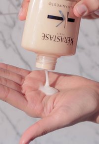 Producto para el cabello Kérastase en una botella de color beige claro, vertiendo una textura cremosa y blanca sobre una palma, mostrando la consistencia del producto.