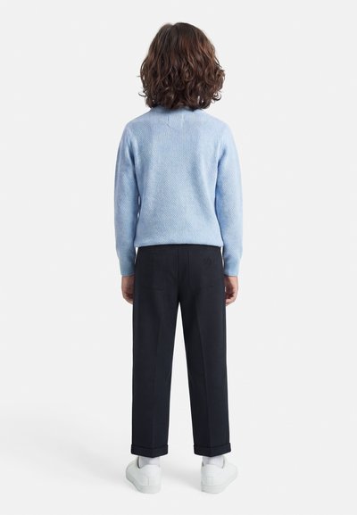 Παιδί με καστανά σγουρά μαλλιά, που φορά ένα ανοιχτό μπλε πουλόβερ, μαύρο παντελόνι, λευκές κάλτσες και λευκά αθλητικά παπούτσια, στέκεται γυρισμένο ανάποδα σε λευκό φόντο.