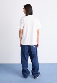 White cotton t-shirt with short sleeves, paired with loose-fitting blue denim jeans featuring a yellow logo patch on the back pocket.
