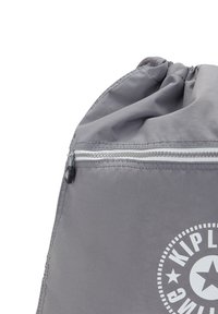 Grå dragsko väska med en slät, glänsande yta. Har en ficka med dragkedja och en cirkulär logotyp med stjärnor i vitt. Kompakt design.