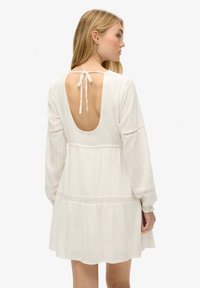 Vestido blanco con mangas largas, espalda al aire y un cierre con lazo. Presenta un borde de encaje y una falda acampanada con una tela texturizada.