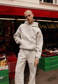 Grå bomulds hættetrøje med en frontlomme og et lille grønt logo, kombineret med matchende sweatpants, der viser en oversized pasform og blød tekstur.
