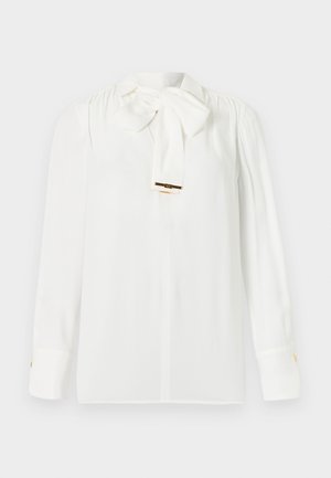 Balta blūze ar garām piedurknēm, lielu sasieto loku pie kakla un zelta pogu detaļām uz aprocēm, attēlota uz vienkārša fona.