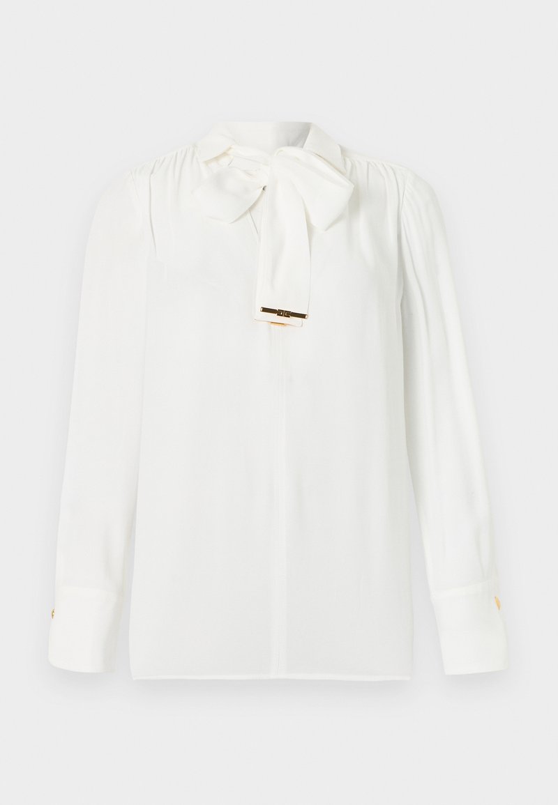Weiße, langärmelige Bluse mit großer, am Hals gebundener Schleife und goldfarbenen Knopfdetails an den Manschetten, dargestellt vor einem einfarbigen Hintergrund.