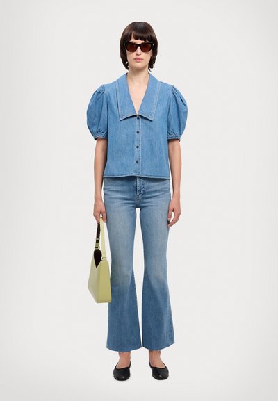 Denim lyhythihainen paita, jossa on suuri kaulus, napitus edessä, yhdistettynä levyileviin farkkuihin; malli pitää kädessään vaaleanvihreää käsilaukkua, päällään tummat aurinkolasit.