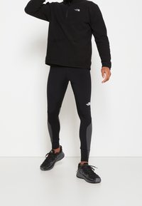 Pullover in pile nero con zip a un quarto, abbinato a leggins neri aderenti con pannelli grigi. Scarpe da ginnastica nere completano l'outfit.