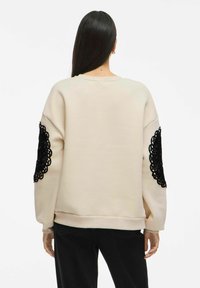 Beige sweatshirt with black lace accents on sleeves, textured fabric, relaxed fit, ribbed cuffs and hem, round neckline, minimalistic design.