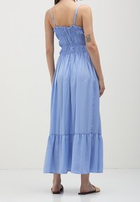 Robe maxi bleue avec corsage froncé, fines bretelles et jupe à volants. Tissu doux, léger avec une texture lisse. Sandales plates portées.