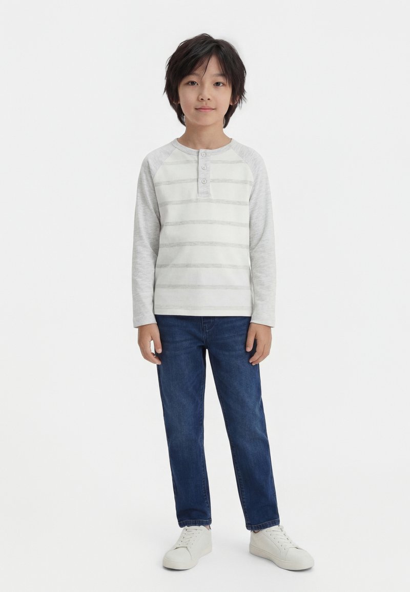 Αγόρι με σκούρα μαλλιά που φοράει ανοιχτό γκρι ριγέ μακρυμάνικο πουκάμισο, μπλε τζιν και λευκά αθλητικά παπούτσια, στέκεται με το πρόσωπο προς τα εμπρός πάνω σε λευκό φόντο.