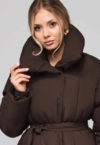 Cappotto imbottito marrone con collo alto, vita con cintura e texture trapuntata. Presenta ampie maniche e una finitura liscia e lucida.