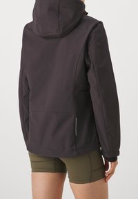 Veste imperméable noire avec capuche, dotée d'une texture lisse, de poches latérales et d'accents réfléchissants. Portée avec un short vert olive.