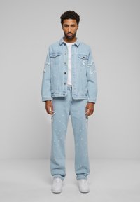 Light blue denim jacket with star patches and distressed details, paired with matching joggers featuring paint splatters. White sneakers complete the look.