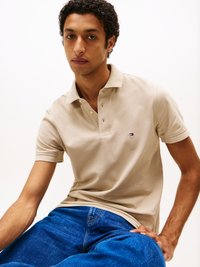 Beige poloing fehér gallérral és három gombbal, amelyen egy kis logó látható. A modellen kék farmernadrág van, amely fehér háttér előtt ül.