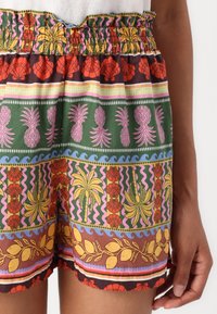 Krótkie spodenki z nadrukiem w kolorowe wzory, przedstawiające ananasy, muszelki, palmy i cytryny, noszone na talii i udzie osoby.