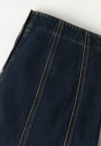 Σκούρο μπλε ύφασμα τζιν με κάθετες πορτοκαλί ραφές και εμφανές μεταλλικό φερμουάρ στο πλάι σε λευκό φόντο.