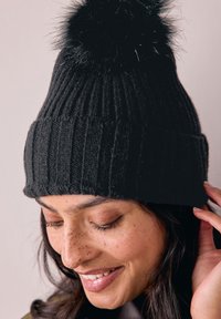 Bonnet en tricot côtelé noir avec un pompon noir moelleux sur le dessus. La texture semble douce, et il épouse parfaitement la tête.
