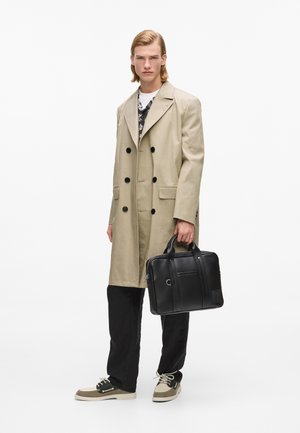 Mladi moški v bež dvovrstnem plašču, črnih hlačah in dvoslojnih supergah, ki drži črno usnjeno aktovko na enostavni belem ozadju.