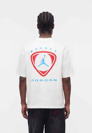 Mann mit geflochtenem Haar, der ein weißes Jordan-T-Shirt mit rotem und blauem Logo und schwarze Hose trägt, von hinten vor neutralem Hintergrund gezeigt.