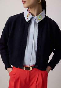 Femme portant un cardigan bleu marine à boutons, une chemise bleu clair avec un col brodé coloré, un pantalon rouge et une ceinture noire avec une boucle dorée.