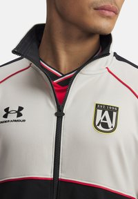 Witte en zwarte zip-up jas met rode accenten. Beschikt over het Under Armour-logo en een schildembleem met "EST. 1996." Gestructureerde stof, atletische pasvorm.