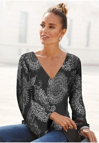 Schwarzes Langarmshirt mit V-Ausschnitt und weißem, kunstvollem Paisleymuster, aus weichem Material, kombiniert mit blauen Jeans und Sonnenbrille.