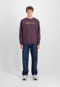 Ljubičasti dukserica s logom i prugastim detaljem na rukavu, uparen s labavim plavim trapericama i sivim tenisicama. Jednostavna pozadina.