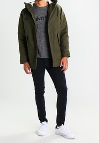 Olivgrön huva parkas över grå crewneck-tröja, kombinerad med svarta jeans och vita sneakers. Minimalistisk design, funktionella fickor, justerbar huva.