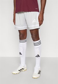 Graue Sportshorts mit weißem Besatz und Adidas-Logo, wei�ße Socken mit schwarzen Streifen und Logo, weiße Fußballschuhe mit schwarzen Akzenten.