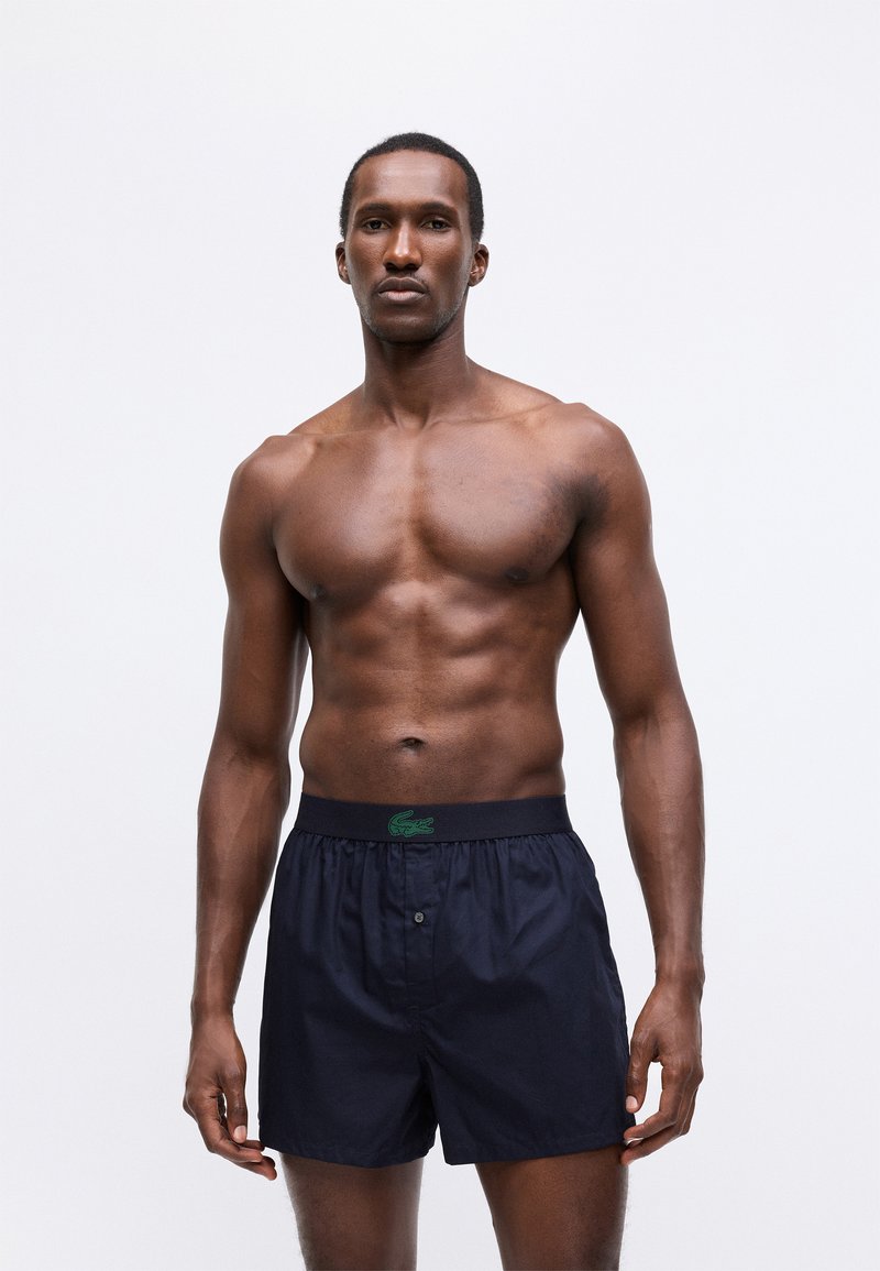 Navy boxer shorts with a green logo waistband, featuring a button closure and a relaxed fit. Smooth fabric with a soft texture.